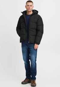 Black padded jacket with a hood, featuring a zip front and visible seams, worn over a navy shirt and denim jeans, paired with brown shoes.