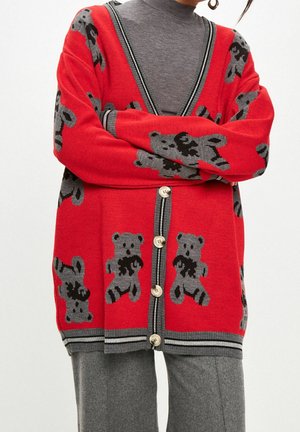 Cardigan rouge avec bordure grise et motifs d'oursons porté sur un col roulé gris et un pantalon gris assorti, bras croisés.