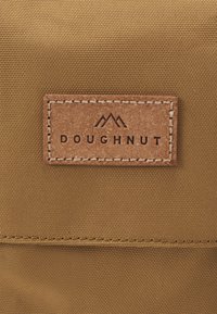 Καφέ υφασμάτινη τσάντα με υφή. Διαθέτει ένα ορθογώνιο δερμάτινο ετικέτα με ανάγλυφο το "DOUGHNUT" και δύο εικονίδια βουνών.