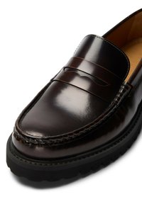 Mocassin en cuir marron au fini brillant, avec un détail de couture surélevée et une semelle en caoutchouc texturée. Dispose d'une bride décorative traversant le cou-de-pied.