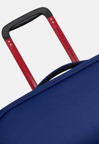 Valigia a rotelle blu con texture, dotata di un manico telescopico nero e rosso, caratterizzato da un'impugnatura morbida e dettagli hardware visibili.