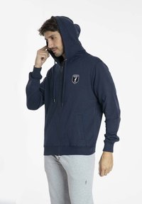 Hoodie zippé bleu marine avec une poche avant et une capuche à cordon, doté d'un patch logo sur la poitrine et de poignets et un ourlet côtelés.