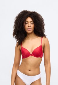 Sarkani polsterēta krūštura ar regulējamām lencēm un loku detaļu starp kausiņiem; ar gludu tekstūru un vieglu mirdzumu. Balti bikini apakšdaļa.
