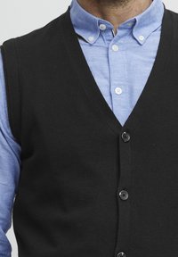 Svart stickad väst med V-halsdesign, prydd med sex knappar och bärs över en ljusblå skjorta med knappar i vitt.