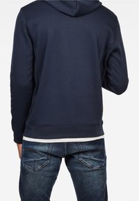 Felpa blu navy con finitura testurizzata, dotata di cappuccio con cordoncino e polsini a coste, abbinata a jeans blu sbiaditi con dettagli sulle tasche.