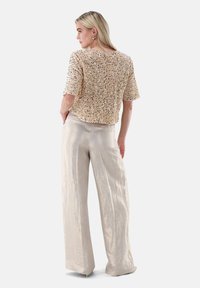 Kurzärmeliges beiges Oberteil mit Pailletten verziert, kombiniert mit hellbeigen Hosen mit weitem Bein und einer glatten, strukturierten Oberfläche.