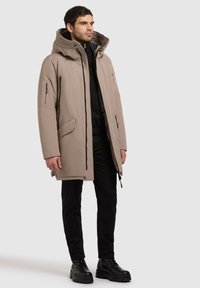 Beige puffatakki korkealla kauluksella, kaksinkertainen vetoketju ja napitus, sivutaskut ja teksturoitu huppu, yhdistettynä mustiin housuihin ja saappaisiin.