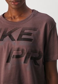 Bordeaux lyhythihainen t-paita, jossa on suuri harmaa lohkoteksti "NIKE" vaakaraidoilla ja pehmeää puuvillamateriaalia.