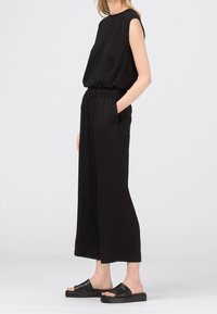 Femme portant un haut noir sans manches, un pantalon noir à jambes larges avec taille élastique et des sandales noires, se tenant de profil avec une main dans la poche.