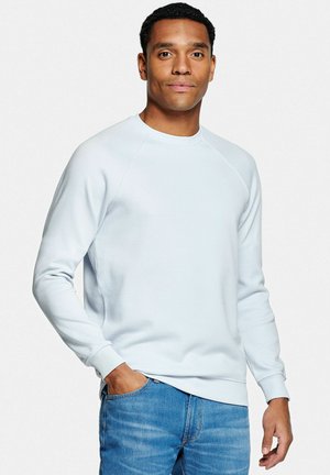 Homme portant un sweat-shirt bleu clair à manches longues et un jean bleu, debout avec une main dans la poche devant un fond uni.