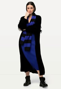 Cardigan nero lavorato a maglia con frange, sopra un vestito blu. Sciarpa a righe nere e blu. Stivali di combat neri con suole spesse.