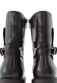 Botas de tobillo de cuero negro con un acabado brillante, que presentan dos cremalleras laterales y correas decorativas con hebillas. Detalle de logo embossado en la parte posterior.