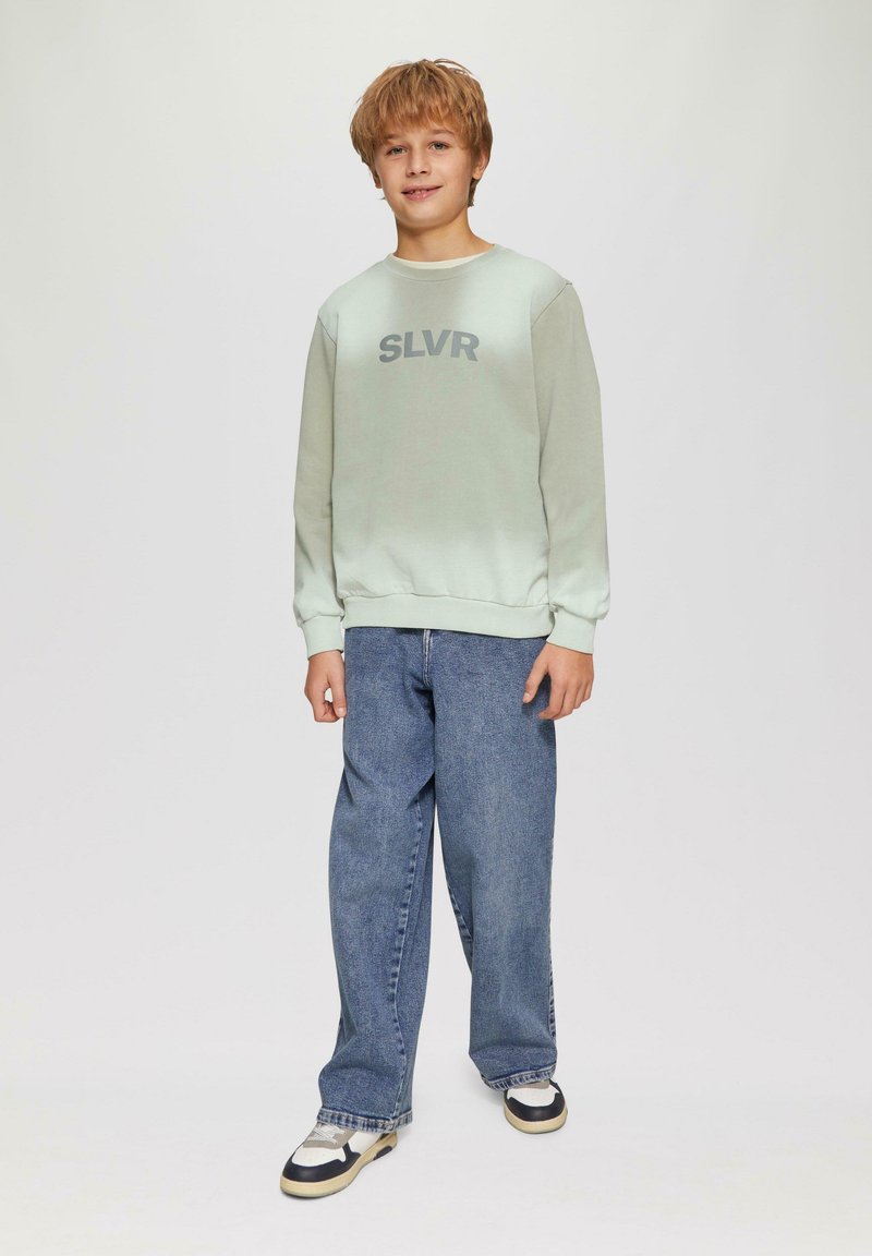 Muntgroene sweatshirt met tekst "SLVR", gecombineerd met losse blauwe jeans en zwart-witte sneakers. Zachte stof en casual pasvorm.