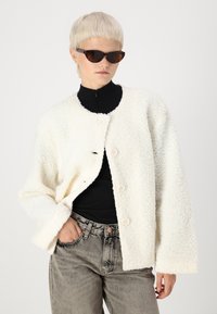 Veste en polaire blanche et moelleuse avec des épaules tombantes, un col rond et quatre boutons. Portée sur un col roulé noir, associée à un jean gris.
