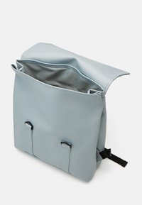 Sac à dos bleu clair avec un rabat ouvert, montrant un intérieur en tissu gris spacieux et deux sangles à boucle noire à l’avant.