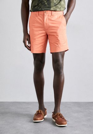 Pomarańczowe bawełniane szorty z płaskim przodem, bocznymi kieszeniami i zapięciem na guzik, zestawione z brązowymi butami żeglarskimi i zieloną koszulą w nadruk.