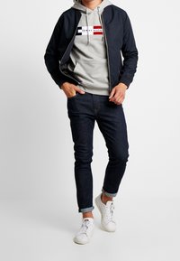 Námorníková bombera s šedou mikinou s logem, doplněná o tmavé džíny a bílé tenisky. Oblečení má slim-fit střih a ležérní styl.