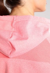 Frau mit einem nackten Hals und dunklen Haaren, die nach hinten gebunden sind, trägt einen rosa Kapuzenpullover mit strukturierten Stoffdetails auf der Rückseite.