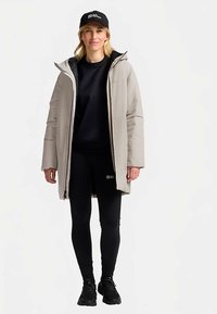 Hellgrauer, isolierter Mantel mit Kapuze, schwarzes Innenfutter, kombiniert mit einem schwarzen Sweatshirt, Leggings und Sportschuhen. Schwarze Kappe mit Logo.