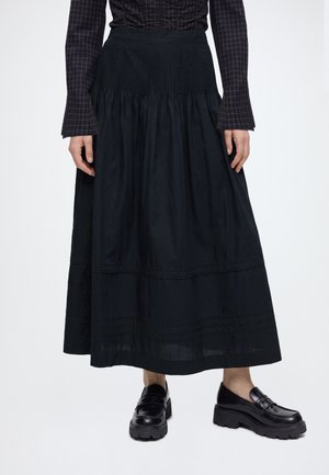 Jupe midi noire à taille froncée portée avec une chemise noire à carreaux à manches longues et des mocassins à plate-forme noirs épais sur fond blanc.
