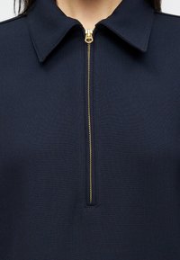 T shirt navy com fecho, com colarinho, apresentando um fecho dourado e uma textura suave, design justo e detalhes de costura mínimos.
