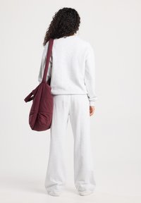 Persona con cabello rizado, vistiendo un chándal gris claro y llevando un gran bolso de hombro acolchado color granate, visto desde atrás sobre un fondo simple.