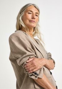 Lys beige blazer med et struktureret ternet mønster, opkrøllede ærmer og sorte knapper. Stof ser blødt og let struktureret ud.