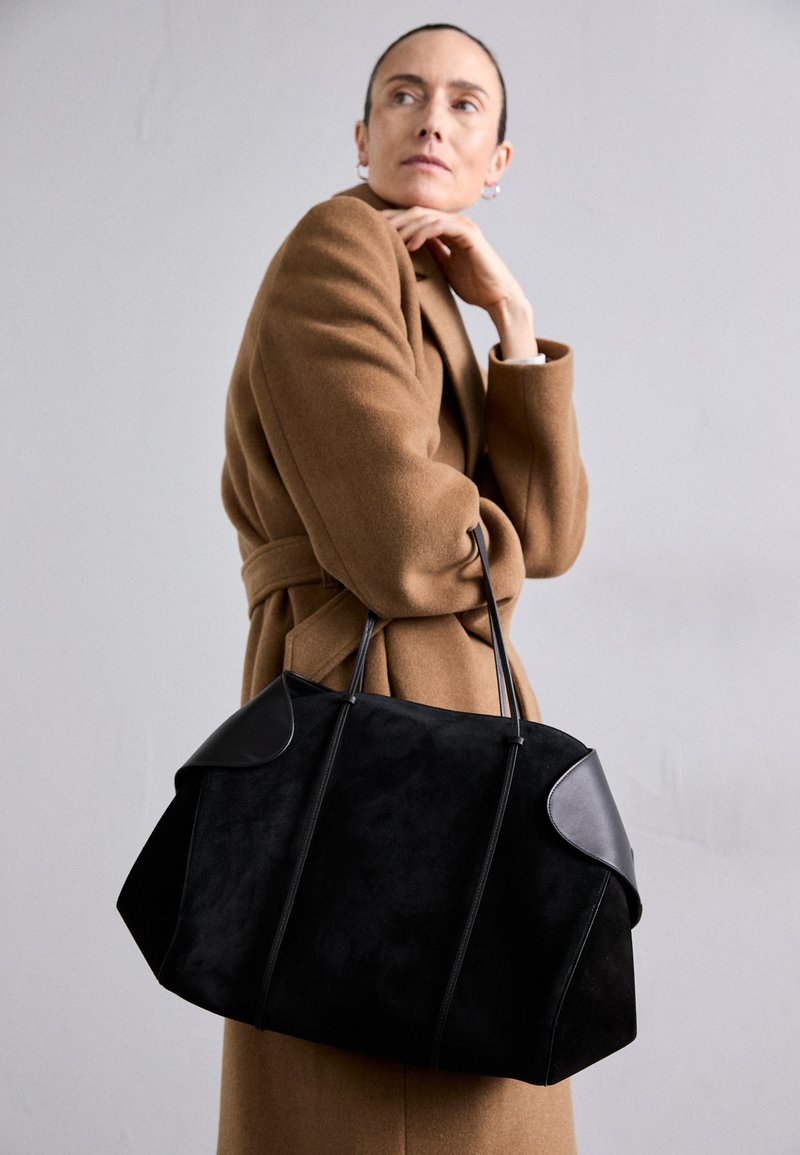 Melna zamšota rokassoma ar ādas akcentiem, strukturēta forma un divas izturīgas rokas, ko nēsā figūra brūnā mētelī.