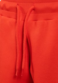 Červené tepláky s žebrovaným pasem. Obsahují stahovací šňůrku s pletenou texturou a hladkou látkou. Detailní záběr na oblast pasu.