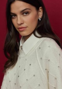 Pull blanc texturé avec des motifs en diamant, orné de petits strass, superposé à une chemise blanche à col.