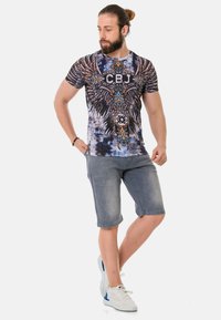 Grafisches T-Shirt mit einem blau-schwarzen Batikmuster, verziert mit Flügeln und komplizierten Designelementen. Kombiniert mit hellgrauen Shorts und weißen Sneakers.