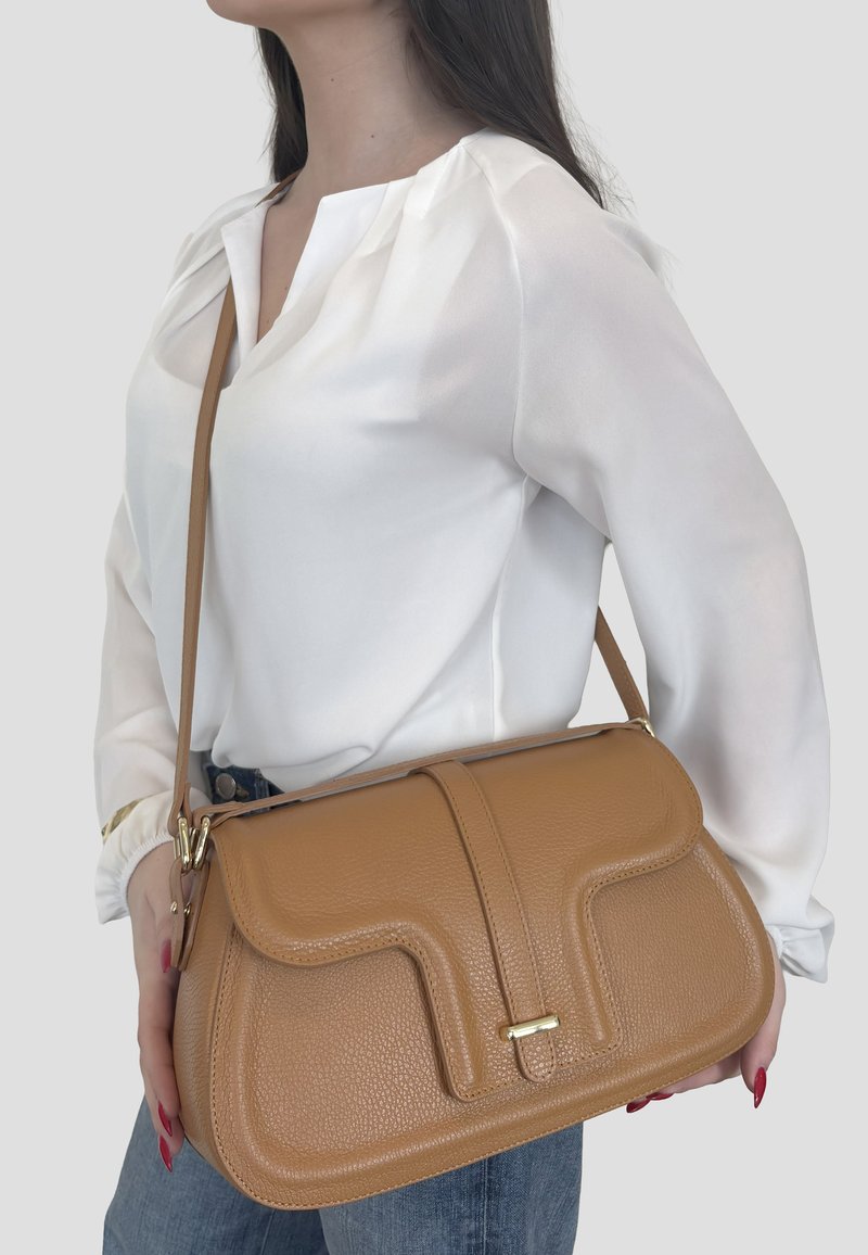 Donna che indossa una camicetta bianca e jeans blu con una grande borsa a tracolla in pelle color cuoio chiaro con dettagli dorati, portata a tracolla.