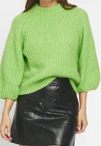 Leuchtend grüner, gestrickter Pullover mit hohem Kragen und puffigen Ärmeln, kombiniert mit einem figurbetonten, schwarzen, kunstledernen Rock mit Krokodilmuster.