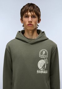 Sudadera color verde oliva con un bolsillo tipo canguro, que presenta tres estampados gráficos en blanco en el lado izquierdo del pecho. Tela suave y texturizada, corte relajado.