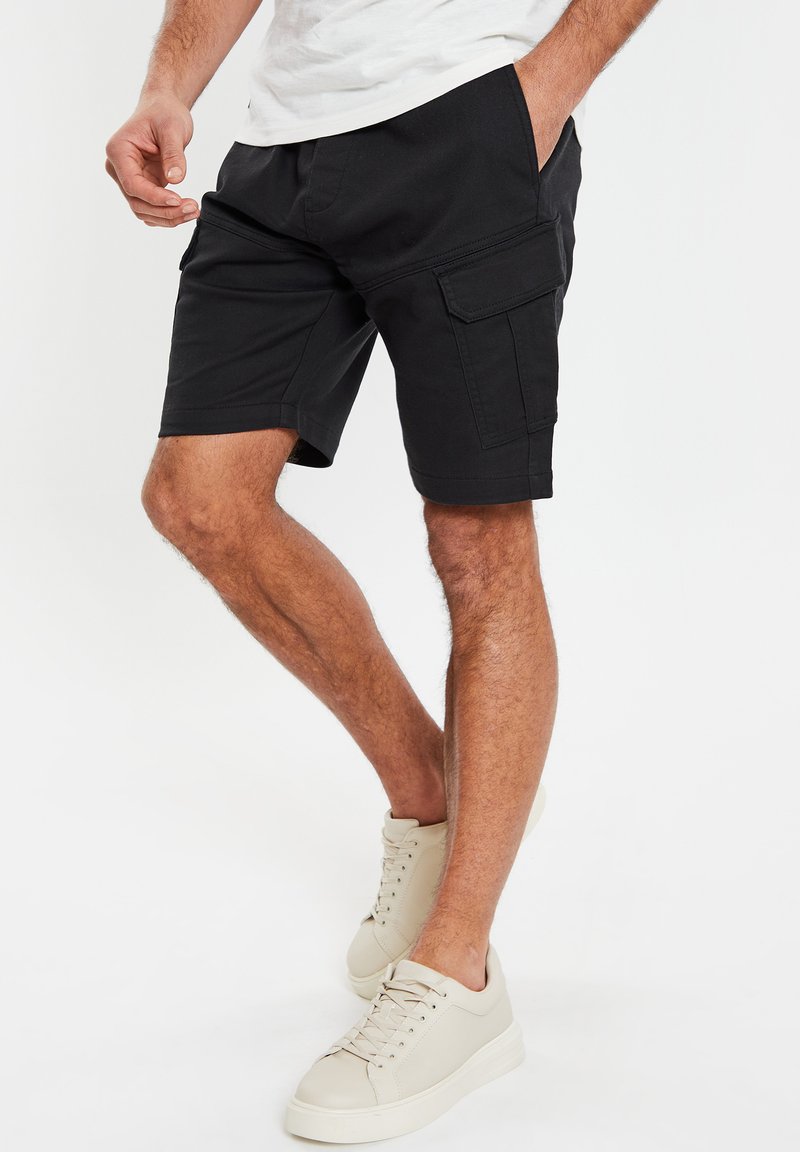 Pantaloni cargo neri con tasche laterali, realizzati in tessuto morbido. Indossati con sneakers chiare, caratterizzate da una silhouette bassa e lacci testurizzati.