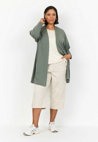 Cardigan verde chiaro sopra un top beige, abbinato a pantaloni cropped beige. Sneakers bianche e grigie completano l'outfit, caratterizzate da dettagli con bottoni.