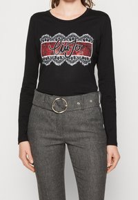 Schwarzes Langarmshirt mit einem dekorativen roten und weißen Grafik, kombiniert mit grauen Hahnentrittmuster-Hosen in Hochbund mit einer runden Gürtelschnalle.