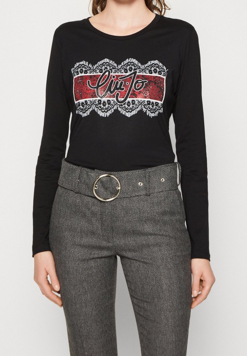 Schwarzes Langarmshirt mit einem dekorativen roten und weißen Grafik, kombiniert mit grauen Hahnentrittmuster-Hosen in Hochbund mit einer runden Gürtelschnalle.