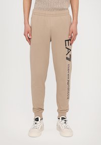 Tan joggebukser med elastisk liv, med sort "EA7" og "Emporio Armani" tekst på siden. Materialet er mykt og glatt.