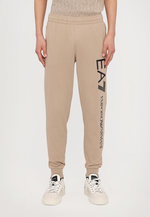 Pantaloni jogger color sabbia con elastico in vita, caratterizzati da testo nero "EA7" e "Emporio Armani" sul lato. Materiale morbido e liscio.