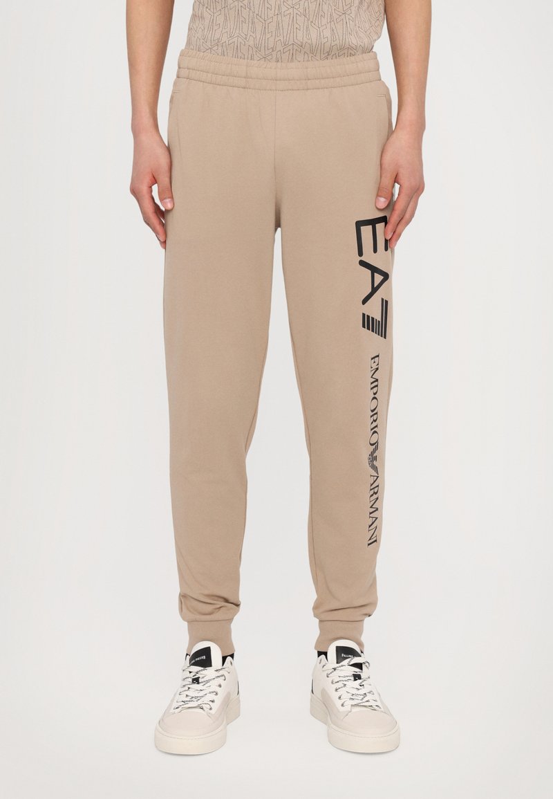 Tan joggebukser med elastisk liv, med sort "EA7" og "Emporio Armani" tekst på siden. Materialet er mykt og glatt.