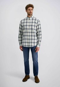 Karierte Button-Down-Bluse in Creme mit Navy- und Grüntönen, kombiniert mit blauen Jeans und braunen Wildlederstiefeln.