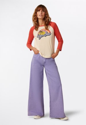 Cremefarbenes und rotes Raglan-Shirt mit einem grafischen Druck, kombiniert mit locker sitzenden, hellvioletten Jeans mit weitem Bein. Schlichtes Design, lässiger Stil.