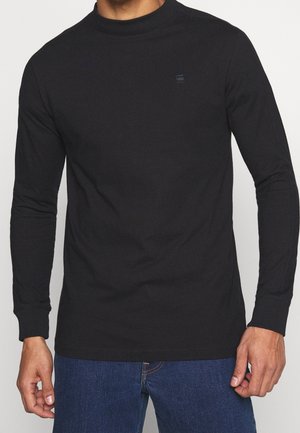 Mand iført en tætsiddende sort langærmet skjorte med et lille logo på brystet, kombineret med blå denimjeans.