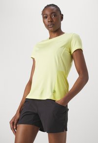 Camiseta deportiva de manga corta, color amarillo claro, con una textura suave y dobladillo curvado, combinada con pantalones cortos negros que cuentan con bolsillos laterales.