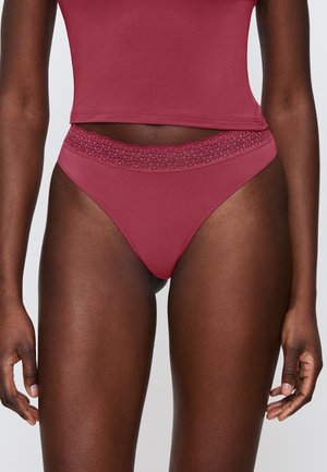 Kobieta ubrana w mauve wysoki strój kąpielowy z koronkowym paskiem i dopasowaną crop top, stojąca na tle białej ściany.