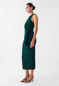 Robe midi sans manches verte avec un corsage ajusté, détail de nouage sur le côté à la taille et texture douce ; associée à des sandales à talons noires.