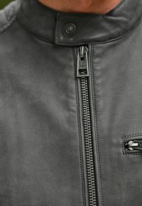 Giacca in pelle nera con colletto alto, zip frontale e materiale texturizzato, dotata di una piccola tasca e dettagli in metallo.