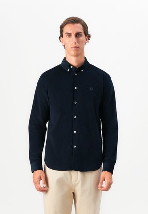 Man with short dark hair wearing a buttoned dark navy shirt and beige pants standing against a plain white background.