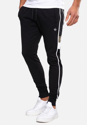 Mann trägt schwarze Jogginghosen mit weißen Streifen an den Seiten und weiße Turnschuhe, steht vor einem einfarbigen Hintergrund.